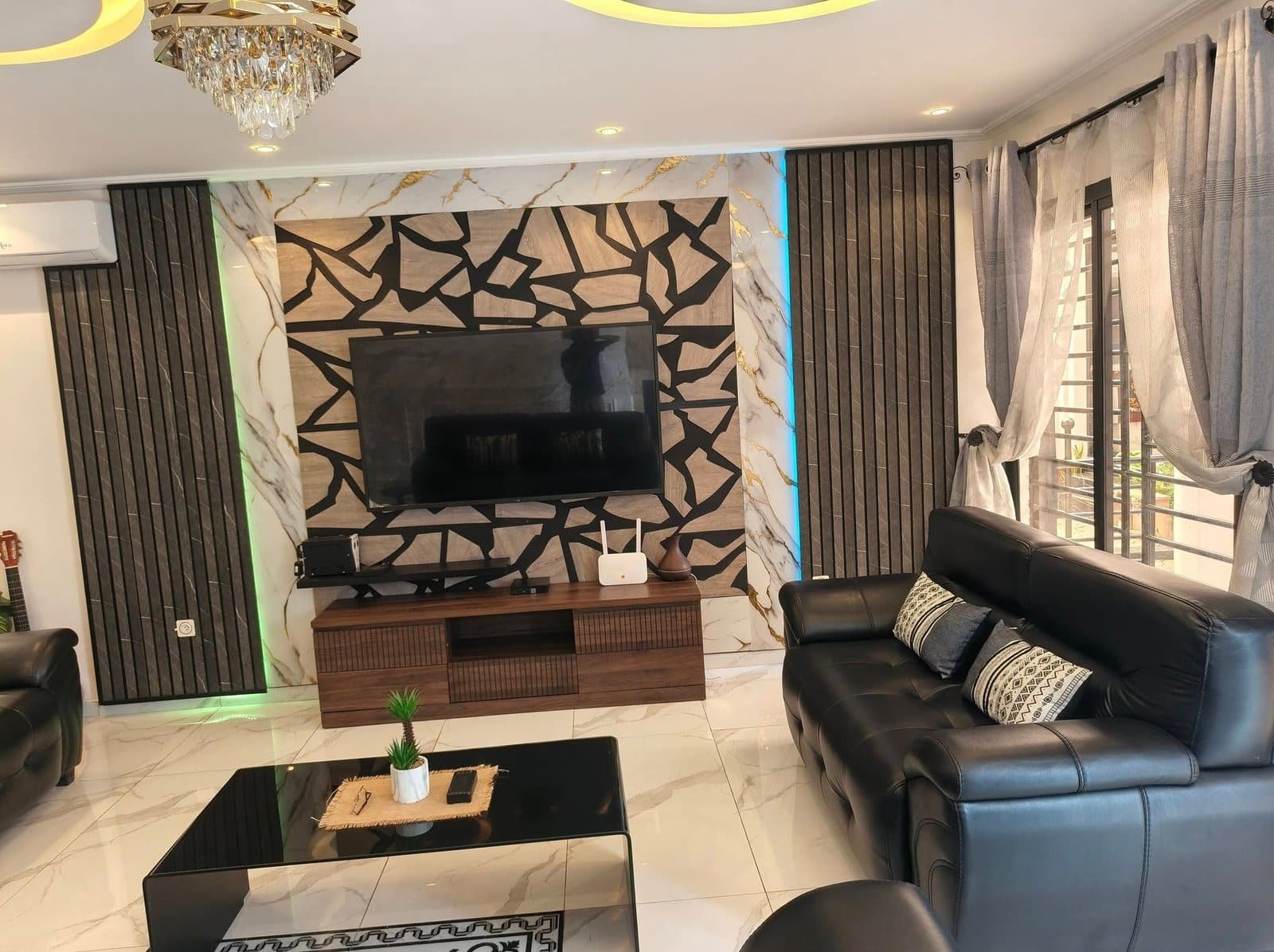 Salon aménagé par NG Design à Douala, avec un mur TV en pierre décorative et bois, des canapés en cuir noir et un sol en marbre